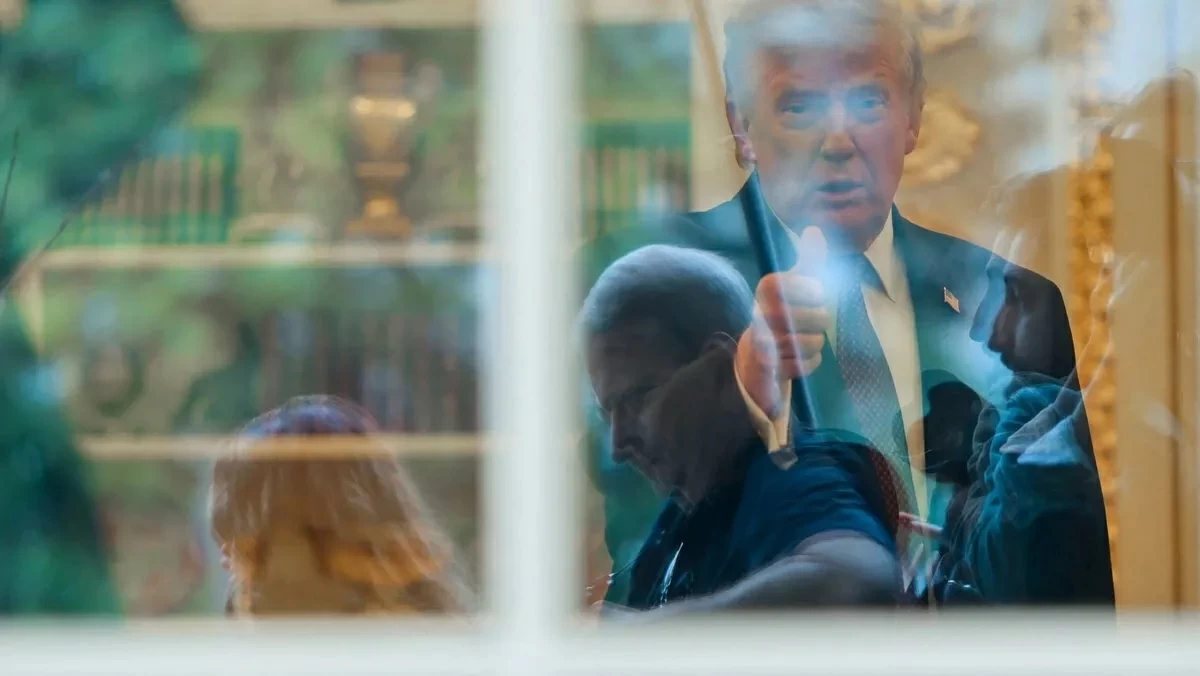 ترمب يلوّح للمصورين من خلف الباب الزجاجي للمكتب البيضاوي.