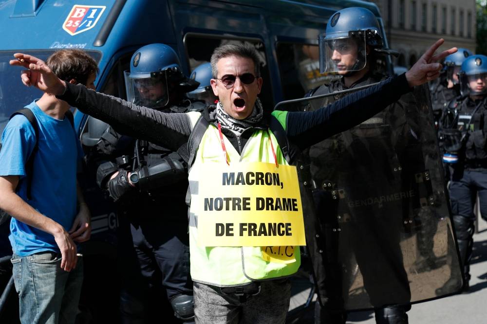 A protester wearing a 'Yellow Vest' (gilet jaune) and sporting a sign reading (a play on word) «Macron, our French drama» in reference to the fire that devastated the Notre-Dame de Paris Cathedral, takes part in an anti-government demonstration in Paris on April 20, 2019. 'Yellow vest' movement protesters take to the streets on April 20, 2019 for 23rd consecutive Saturday. / AFP / Zakaria ABDELKAFI 