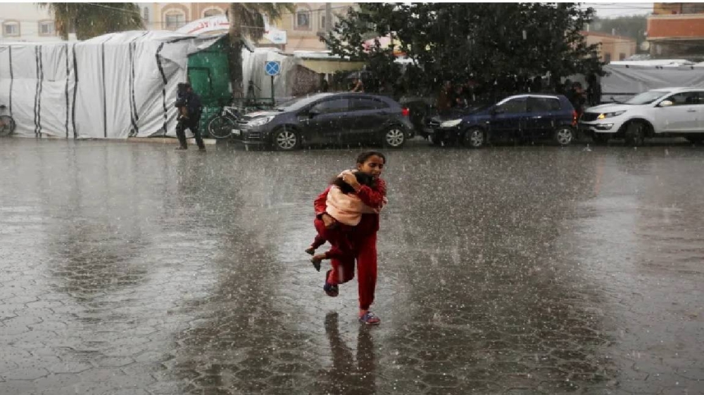 طفلة فلسطينية تحمل شقيقتها في تجسيد لمعاناة النازحين الذين يواجهون البرد والمطر والقصف الإسرائيلي .